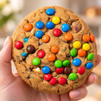 A close-up of a hand holding a large chocolate chip cookie topped with colorful M&M's candies, with a softly blurred indoor background.